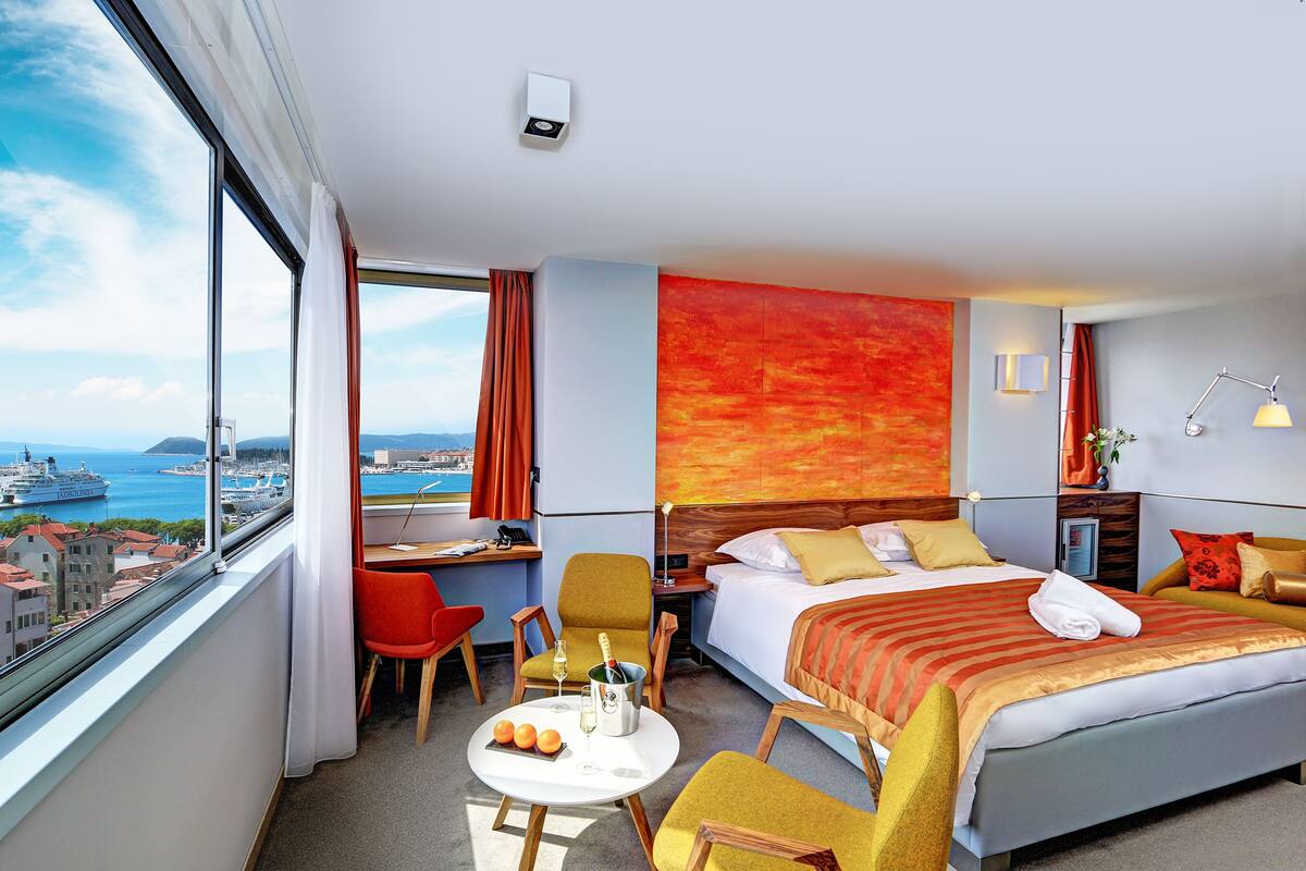 A hotel room with a large bed, a desk, a chair, a small table with a bottle and glasses, and a view of the sea and buildings.