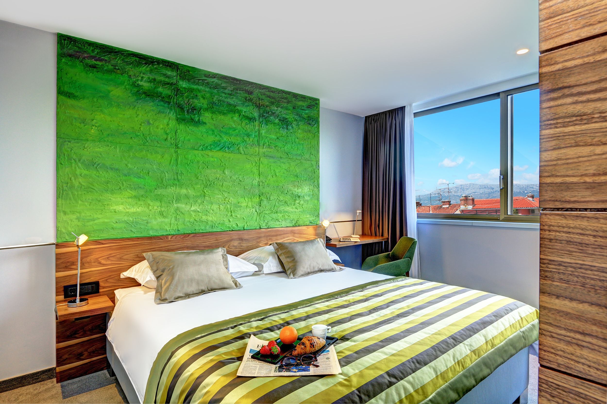 Une chambre d’hôtel avec un grand lit, un mur décoré d’un motif abstrait vert, une vue sur les immeubles par la fenêtre et un plateau de petit-déjeuner posé sur le lit.