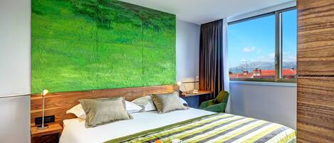 Een hotelkamer met een groot bed, een abstracte groene wand, uitzicht op gebouwen door een raam en een ontbijtkarretje op het bed.