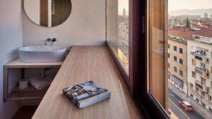 Un baño moderno con un espejo redondo, un lavamanos blanco y un mueble de tocador de madera. Una revista está sobre el mueble y la habitación tiene vistas a un paisaje urbano a través de un ventanal grande.