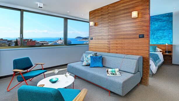 Una habitación de hotel moderna con un ventanal grande, un sofá gris, sillas color turquesa, una mesita redonda y una pared de acento de madera.