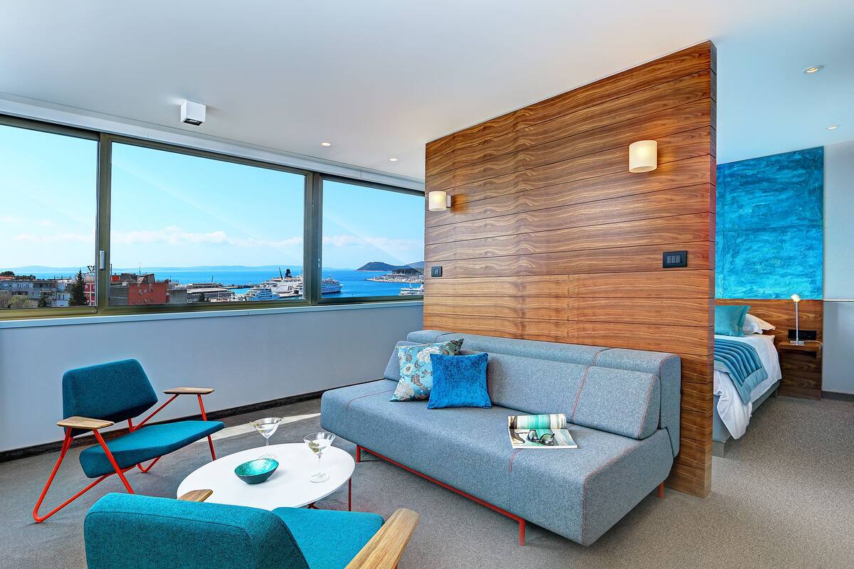A modern hotel room with a large window, a grey sofa, teal chairs, a small round table, and a wooden accent wall.