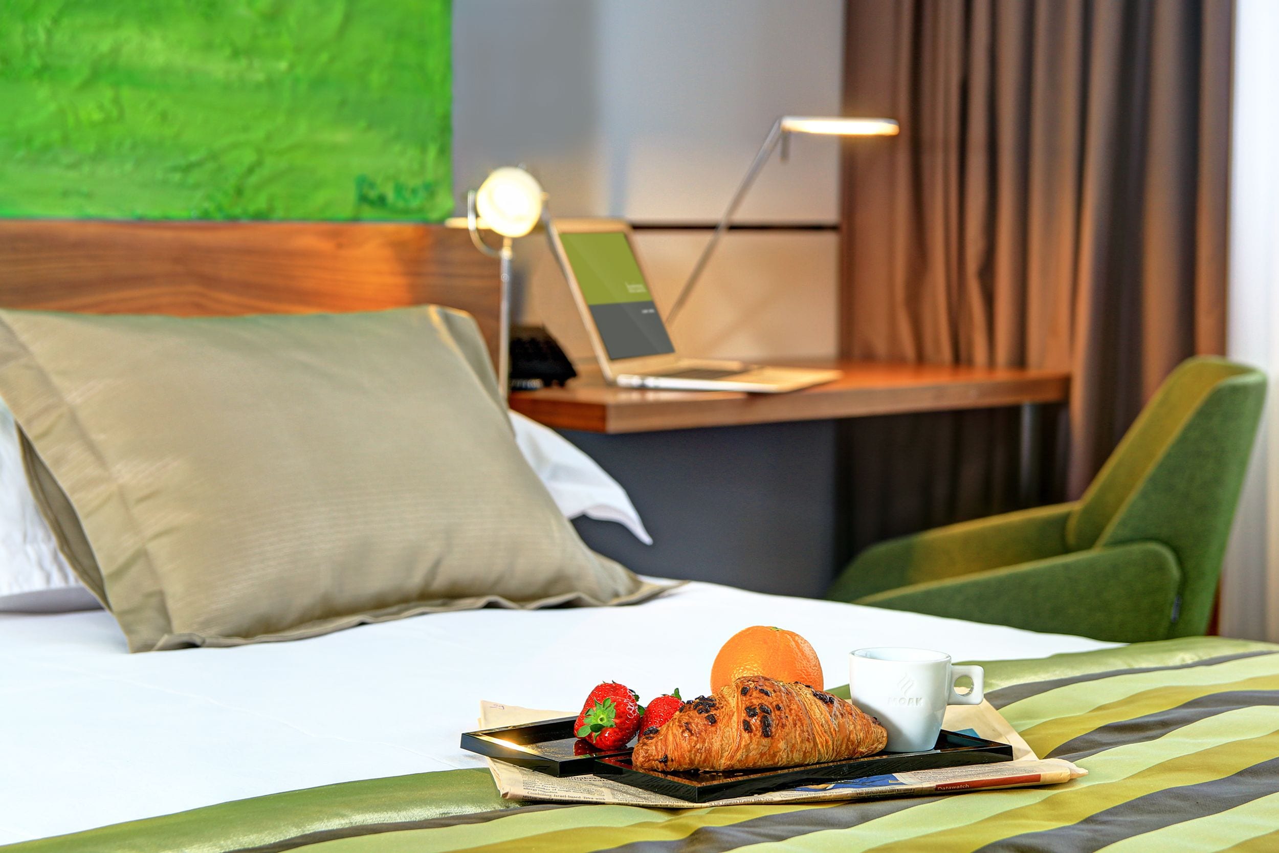 Een net opgemaakt bed met een dienblad ontbijtproducten, een laptop op een bureau en een groene stoel in een moderne kamer.