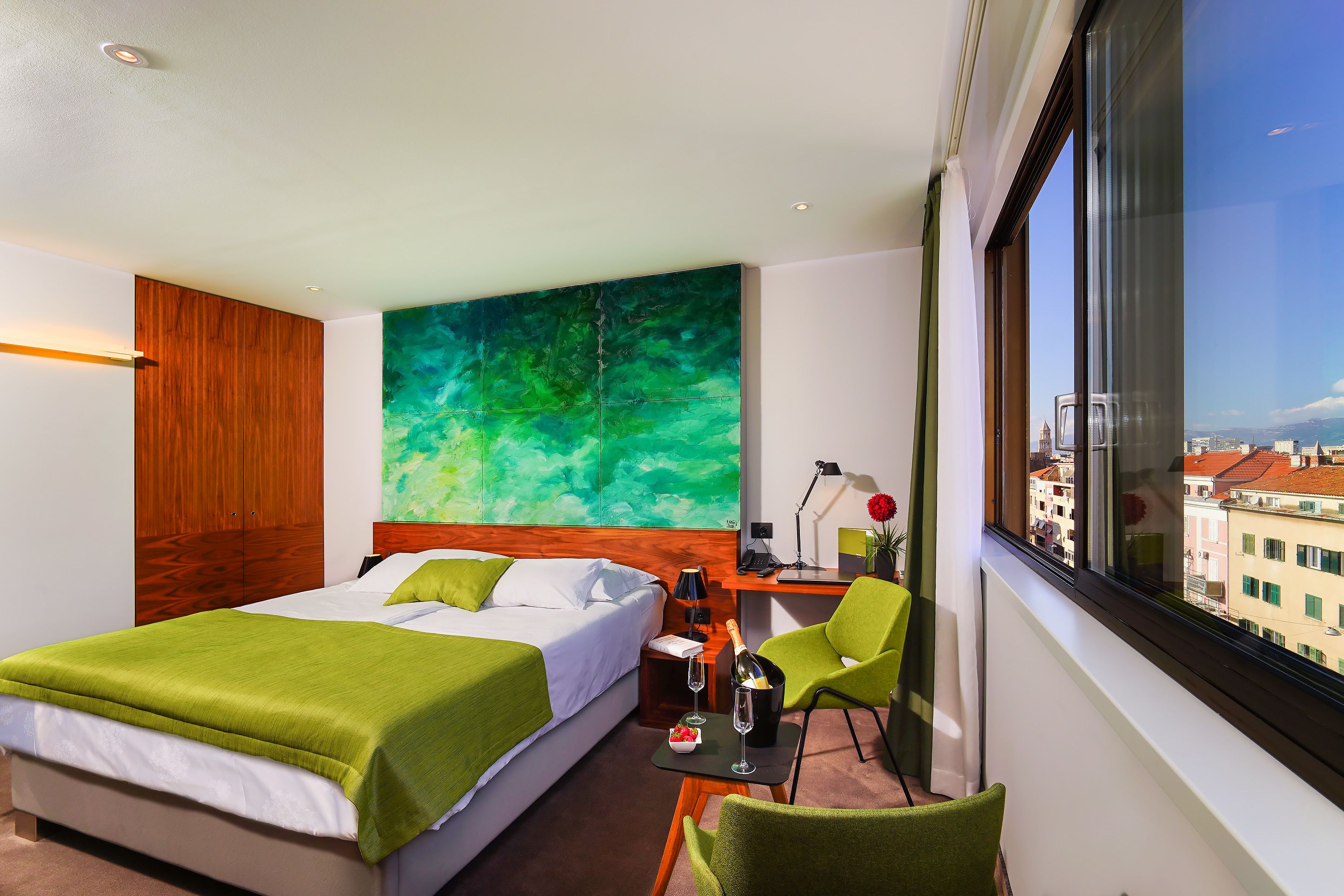 A hotel room with a bed, a desk with a chair, a large window, and a green abstract painting on the wall.