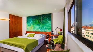 A hotel room with a bed, a desk with a chair, a large window, and a green abstract painting on the wall.