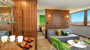 Una habitación de hotel moderna con una pared de acento de madera, un sofá, un área de comedor con una bandeja de croissants y vistas a la ciudad a través de amplias ventanas.