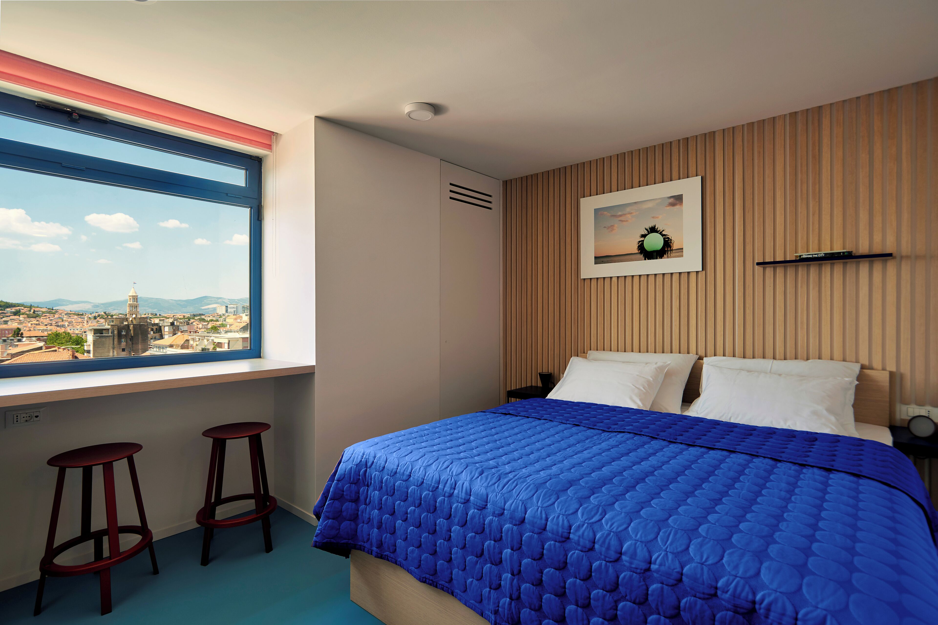 Un dormitorio con un ventanal grande, una cama con colcha azul, dos taburetes rojos y una estantería en la pared.
