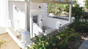 A white building with a covered porch, steps leading to a door, and a garden with flowering plants. - Apartment in Ag.Anna area,fully equipped,semi-basement & very quiet environment. (Νάξος)