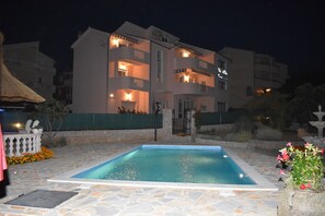 A swimming pool with a stone deck, surrounded by a building with multiple balconies and a well-lit entrance.