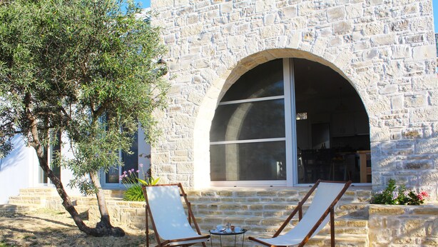 A stone building with a large arched doorway, two white lounge chairs, and a small table with cups and saucers. - Villa Marikami, an amazing new stone built house in Kamilari (Καμηλάρι)