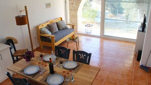 A dining area with a wooden table set for two, a sofa, and a view of the outdoors through a large window. - Villa Marikami, an amazing new stone built house in Kamilari (Καμηλάρι)