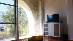A room with a large window, a television on a stand, and a fruit bowl on a glass table. - Villa Marikami, an amazing new stone built house in Kamilari (Καμηλάρι)