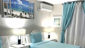 A hotel room with two beds, each with a blue bedspread, white headboards, and bedside lamps. There is a window with a view of palm trees and a beach, and an air conditioning unit mounted on the wall. - Ocho Rios Vacation Resort Property Rent (Ocho Rios)