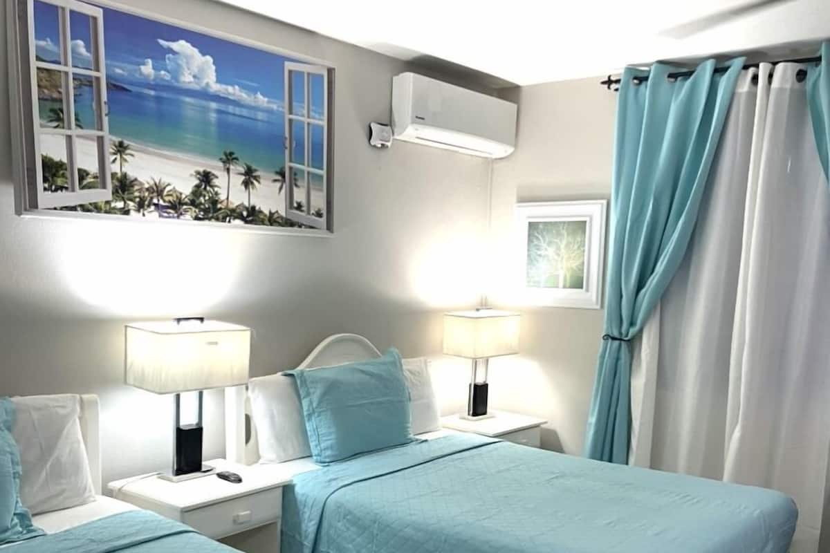 Ett hotellrum med två sängar, var och en med ett blått överkast, vita sänggavlar och sänglampor. Det finns ett fönster med utsikt över palmer och en strand, och en luftkonditioneringsanläggning som är monterad på väggen.