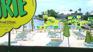 A beachside view with lounge chairs and umbrellas, a sign for 'Land Village' in Ocho Rios, Jamaica, and a body of water in the background. - Ocho Rios Vacation Resort Property Rent (Ocho Rios)