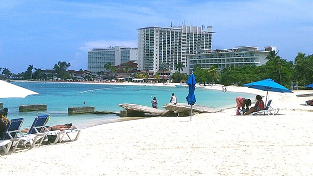 A beach with sun loungers, a pool, and a hotel in the background. - Ocho Rios Vacation Resort Property Rent (Ocho Rios)