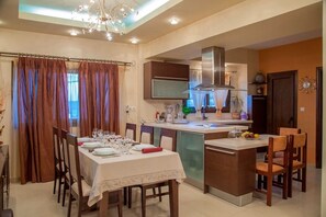 A modern kitchen with a central island, a dining area with a table set for four, and a chandelier. - Villa Veghera - Join Crete, Seaview, private pool (Kissamos)