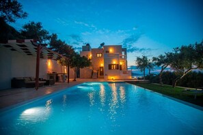 A modern poolside area with a clear view of the pool, a covered seating area, and a building with multiple levels and outdoor lighting. - Villa Veghera - Join Crete, Seaview, private pool (Kissamos)