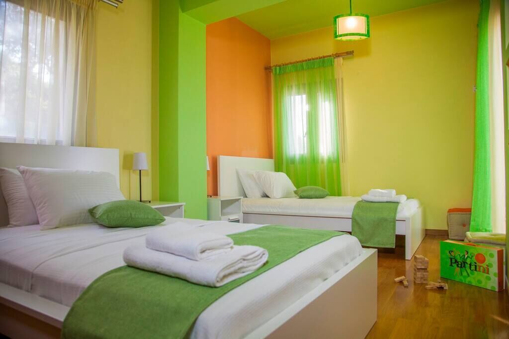 A hotel room with two beds, green and white bedding, a bedside table with a lamp, and a green curtain.