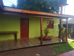 A green building with a tiled porch, a wooden bench, a red door, and potted plants.