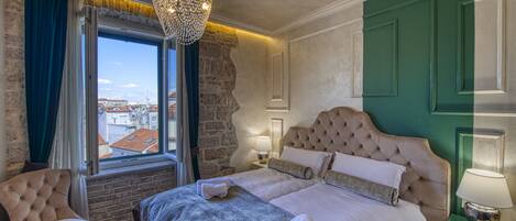 A hotel room with a large bed, a chair, a window with curtains, a chandelier, and a view of buildings outside.