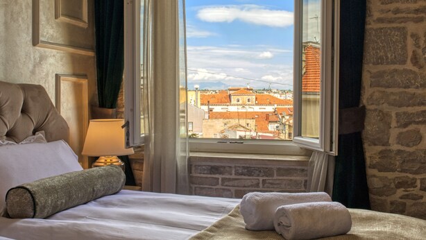 A hotel room with a bed, two rolled towels, a window with curtains, and a view of buildings. - Riva Palace - design rooms (Zadar)