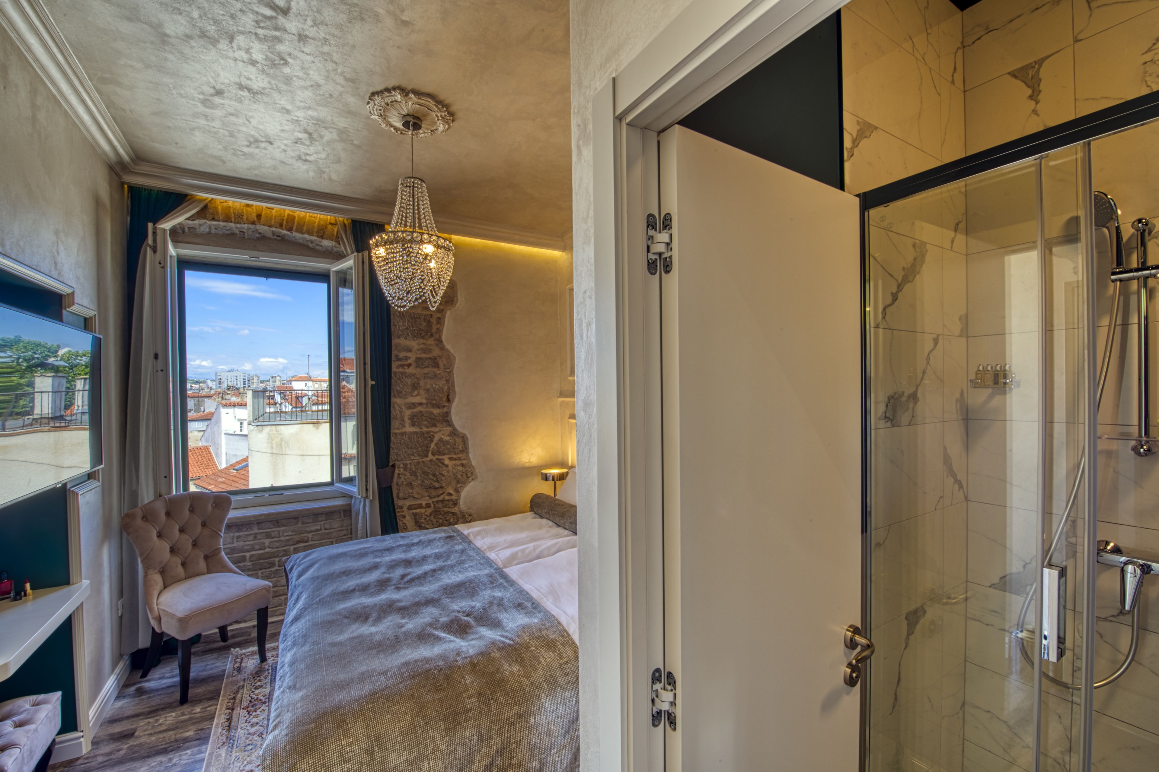 A bedroom with a bed, a chair, a large window, and a shower area.