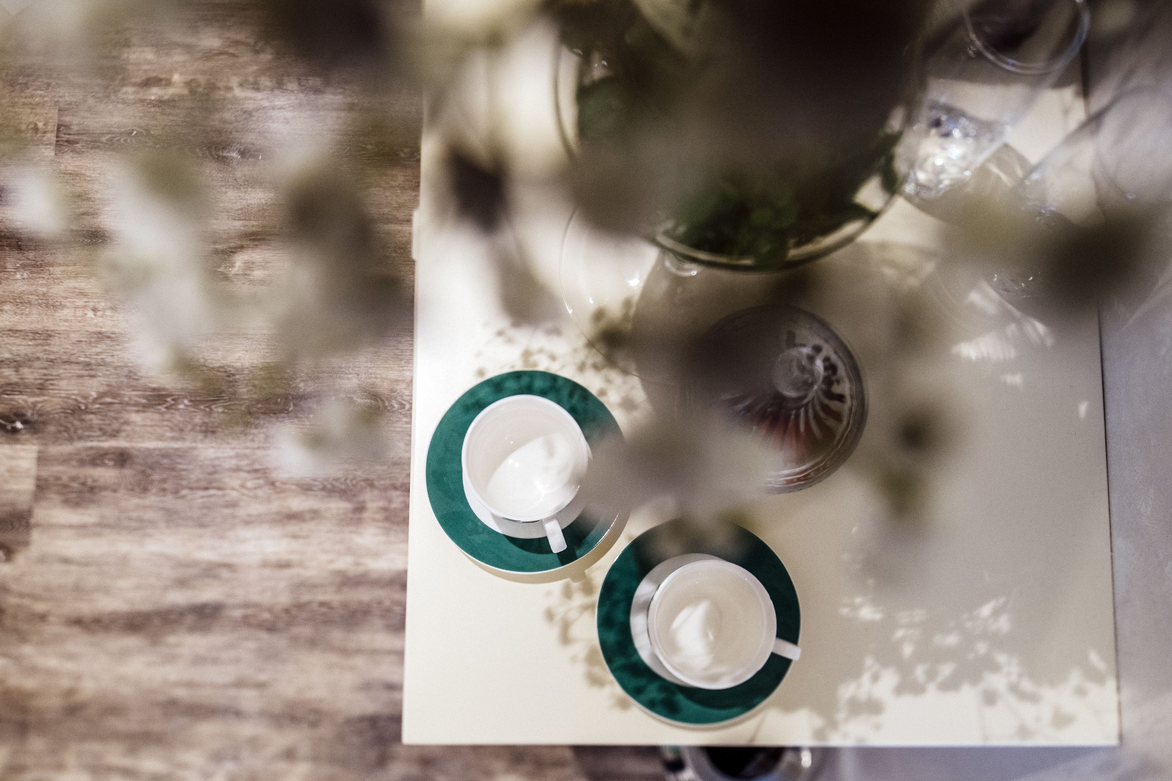 Una mesa con dos tazas blancas sobre platillos verdes, un vaso de agua y un fondo desenfocado.