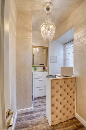A hallway with a white door, a wooden floor, a white desk with a laptop, a mirror, and a chandelier.