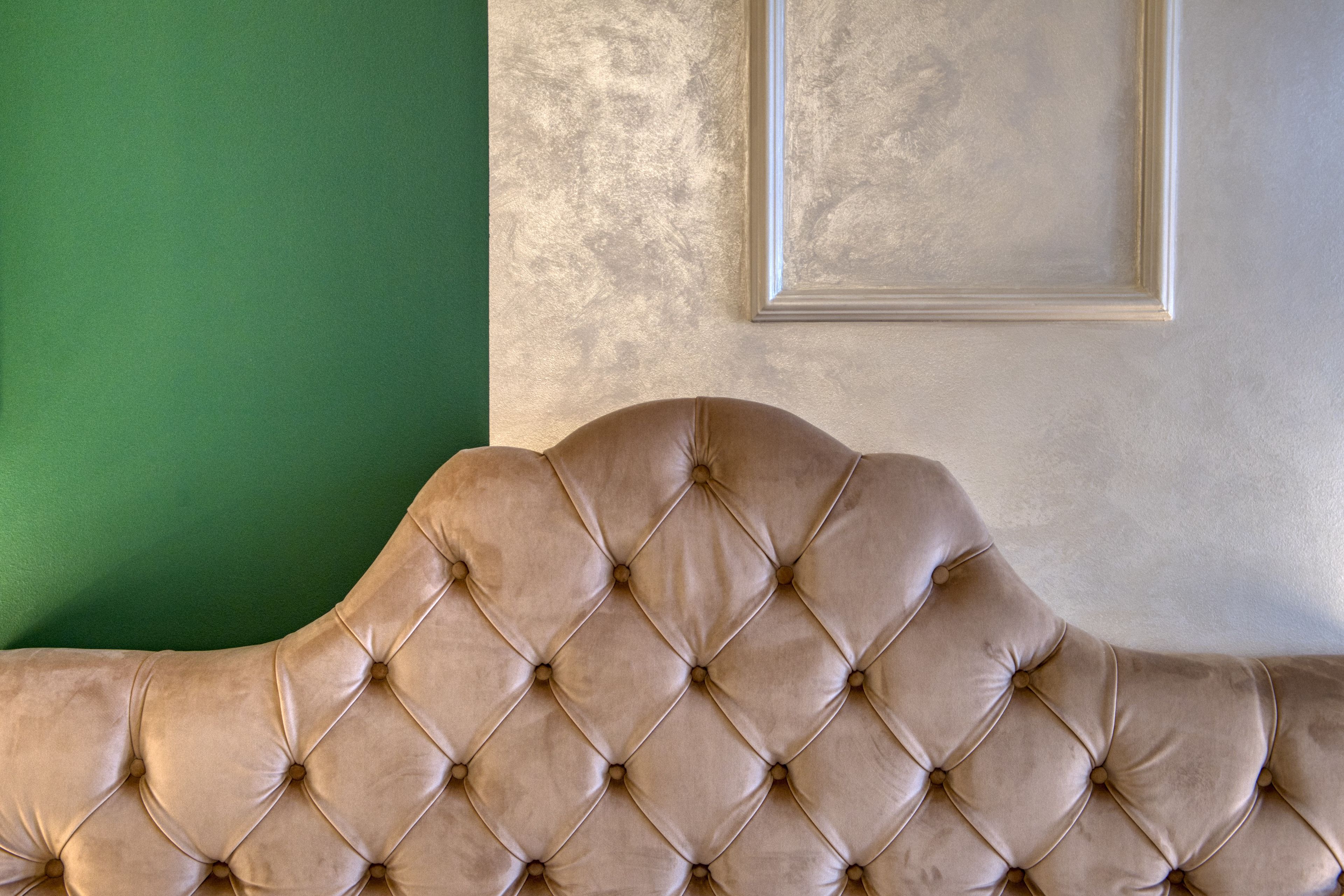 Une tête de lit capitonnée se dresse contre un mur vert, lequel est agrémenté d’un mur texturé de couleur grise et d’un tableau encadré au-dessus.