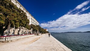 A waterfront promenade with a stone pathway, trees, and a building with columns. - Riva Palace - design rooms (Zadar)