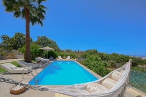 A swimming pool with lounge chairs and a hammock, surrounded by greenery and a clear sky.