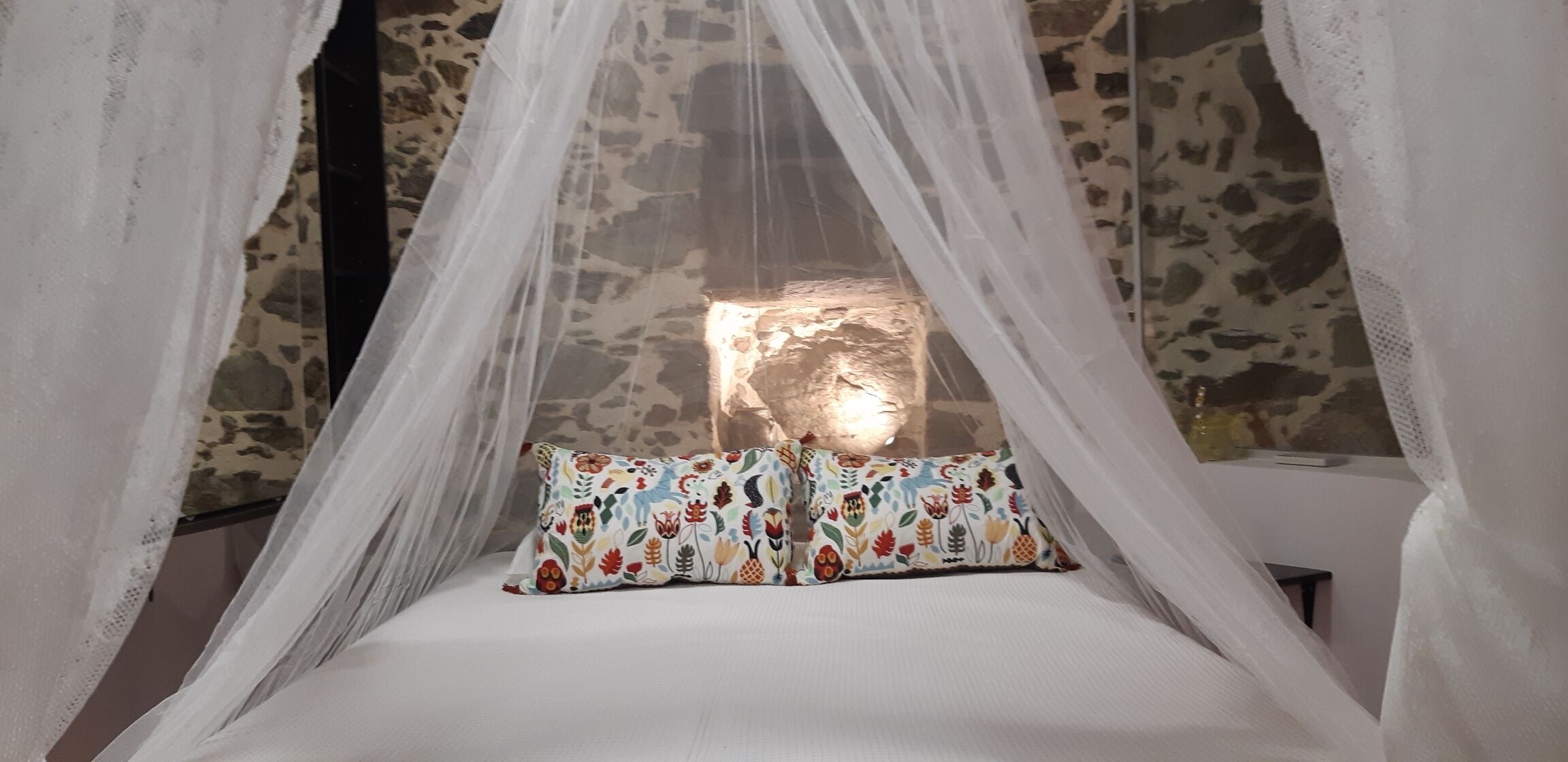 Una cama con dosel, un cojín floral y una pared de piedra al fondo.