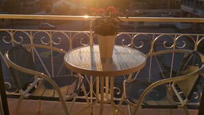 En balkong med ett runt bord och stolar, en krukväxt med blommor och en utsikt över byggnader och den nedgående solen i bakgrunden.