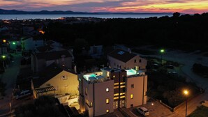 En modern byggnad med en pool på taket och flera balkonger, omgiven av ett bostadsområde med synliga gator och parkerade bilar.