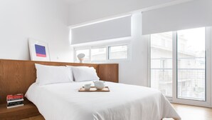 A modern bedroom with a large bed, wooden headboard, and a view of buildings through large windows. - Modern 2 bed/2 bath Acropolis view flat (Αθήνα)