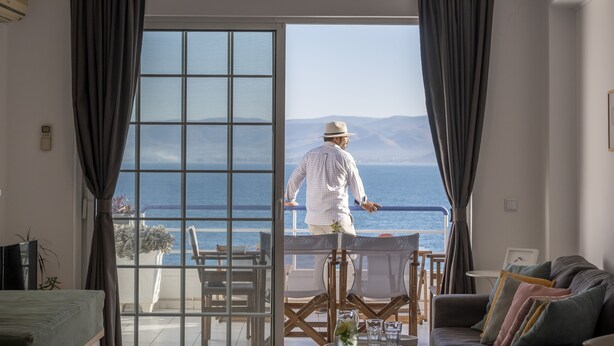 A room with a large sliding glass door leading to a balcony with a view of the sea and mountains.