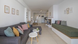 A modern living room with a grey sofa, a green sofa, and a small white table with drinks. There is a kitchen area in the background with a dining table and chairs.
