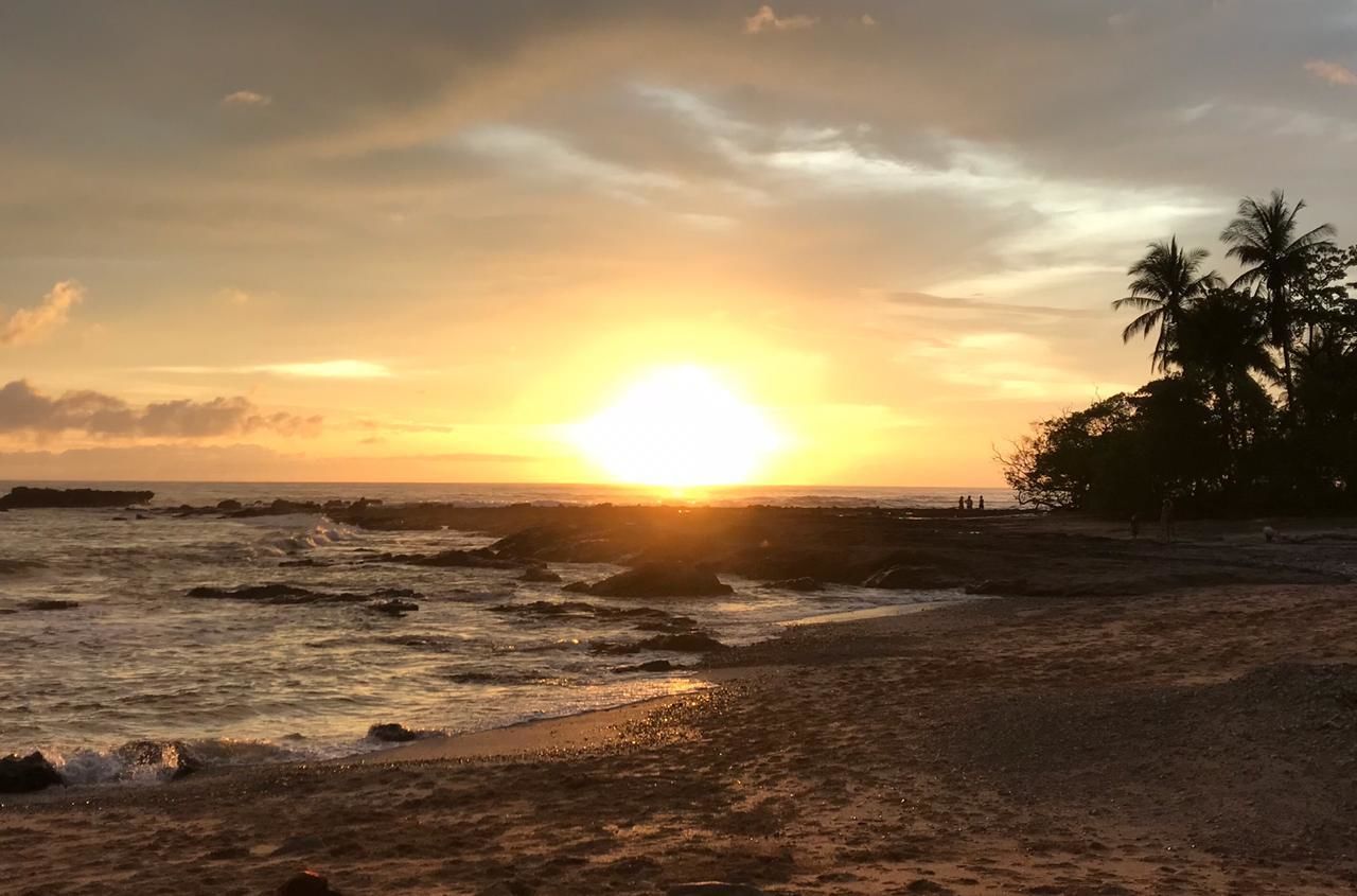 Ein Strand bei Sonnenuntergang, die Sonne tief am Horizont, Palmen und Felsformationen.
