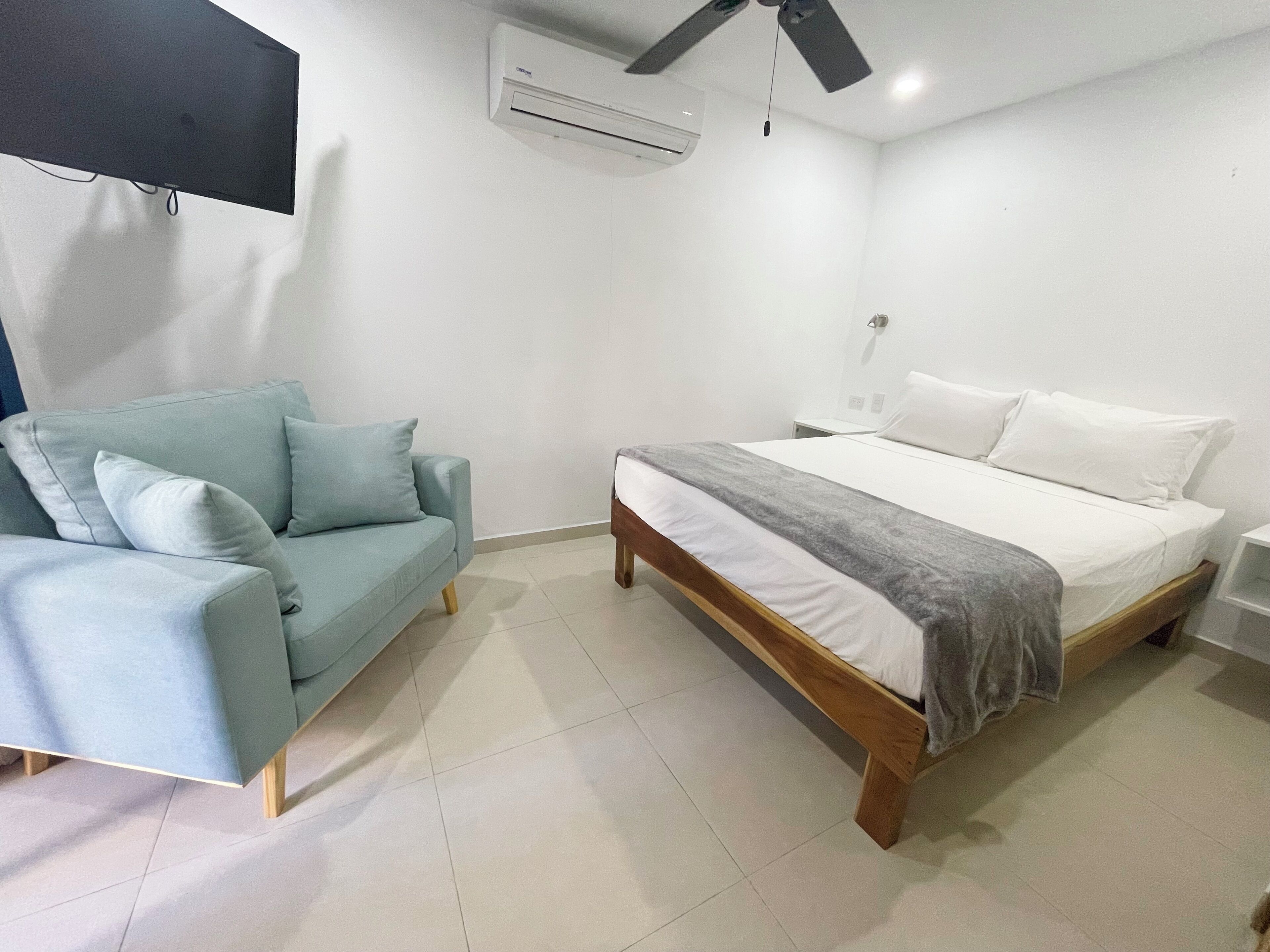 Quarto com cama, sofá, televisão na parede e ar condicionado.