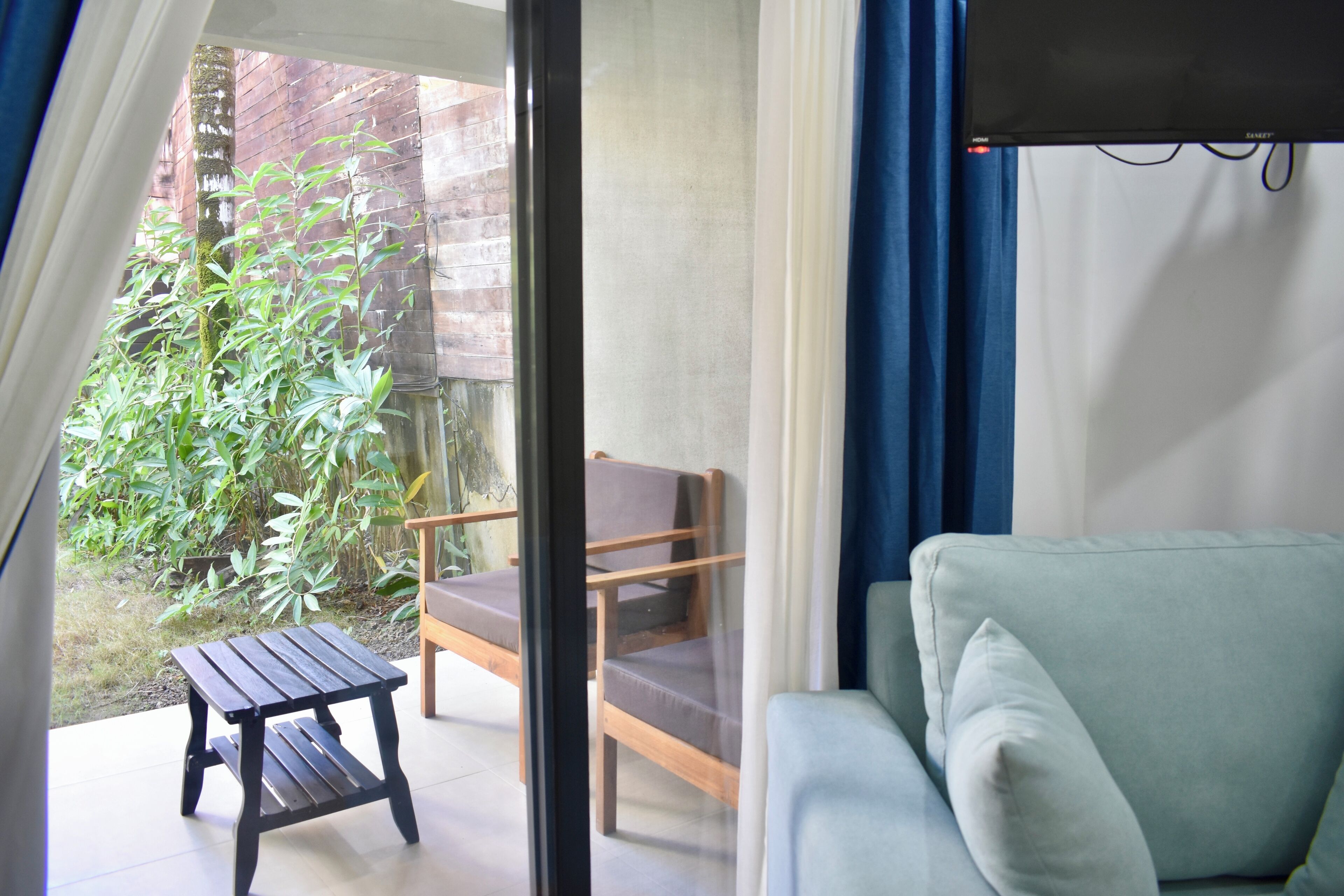 A room with a green sofa, a wooden chair, and a small table. There is a view of a garden with plants and a brick wall.