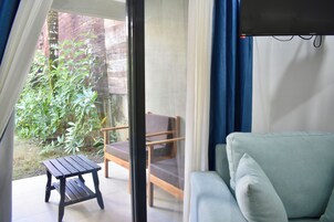 Una habitación con un sofá verde, una silla de madera y una mesita. Se aprecia una vista de un jardín con plantas y un muro de ladrillo.