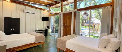 A room with two beds, a desk, and a TV. There is a sliding glass door leading to an outdoor area with a pool and palm trees.