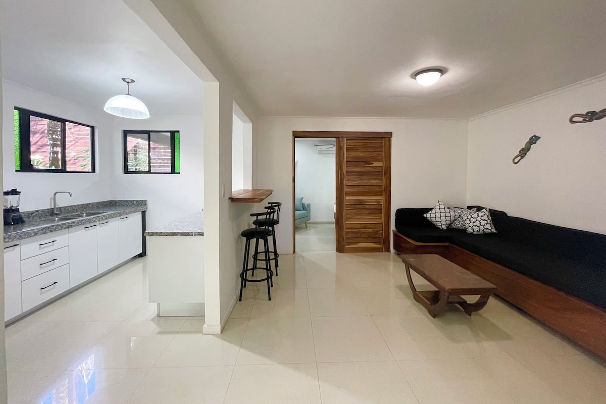Una cocina moderna con armarios blancos y encimera de granito, una mesa de comedor de madera con sillas negras, y un sofá negro con cojines estampados.