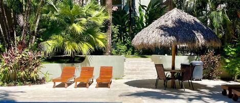 A pool area with lounge chairs and a thatched umbrella, surrounded by lush tropical vegetation.