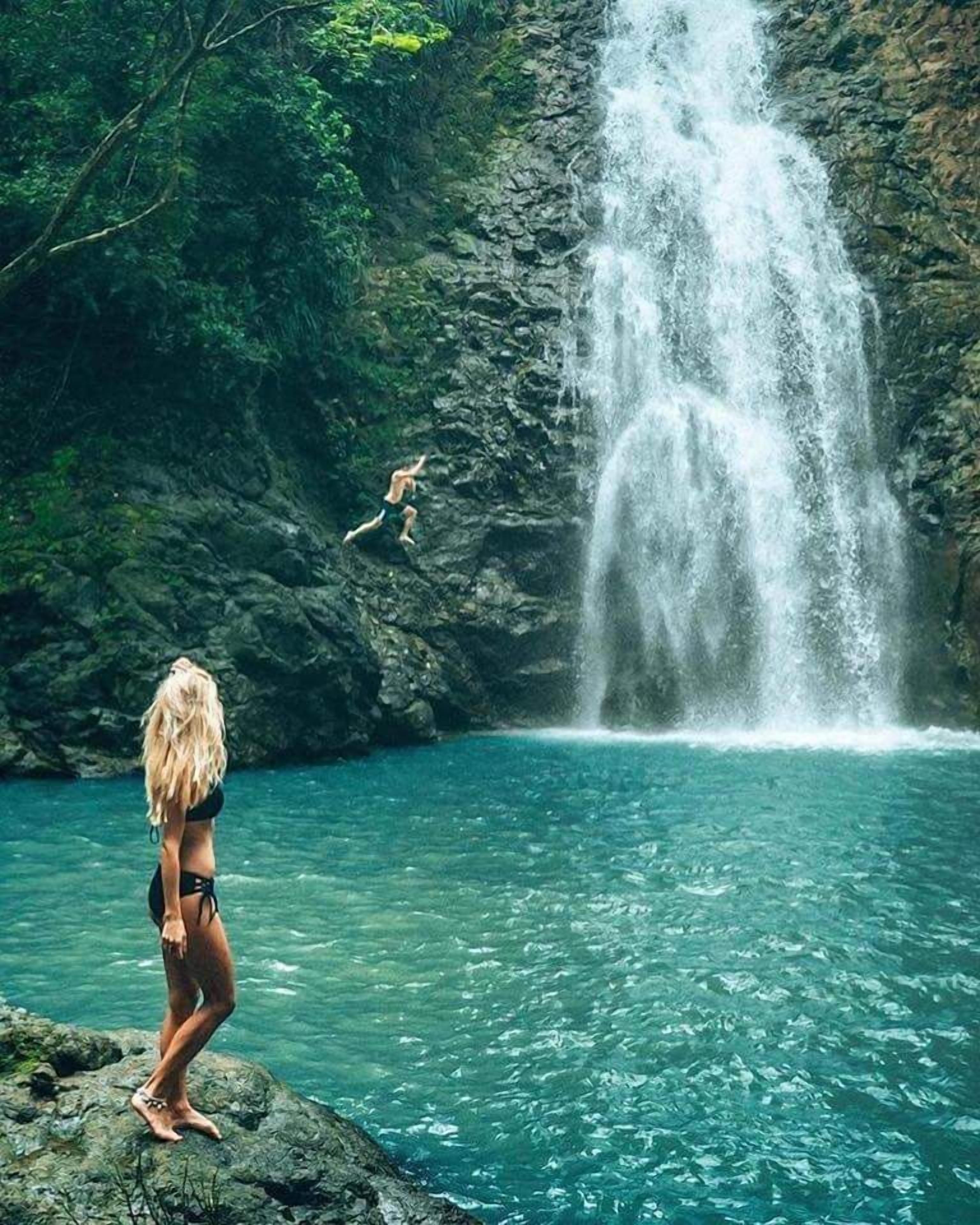 A woman in a bikini standing on rocks by a clear blue pool with a waterfall in the background.