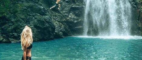 A woman in a bikini standing on rocks by a clear blue pool with a waterfall in the background.