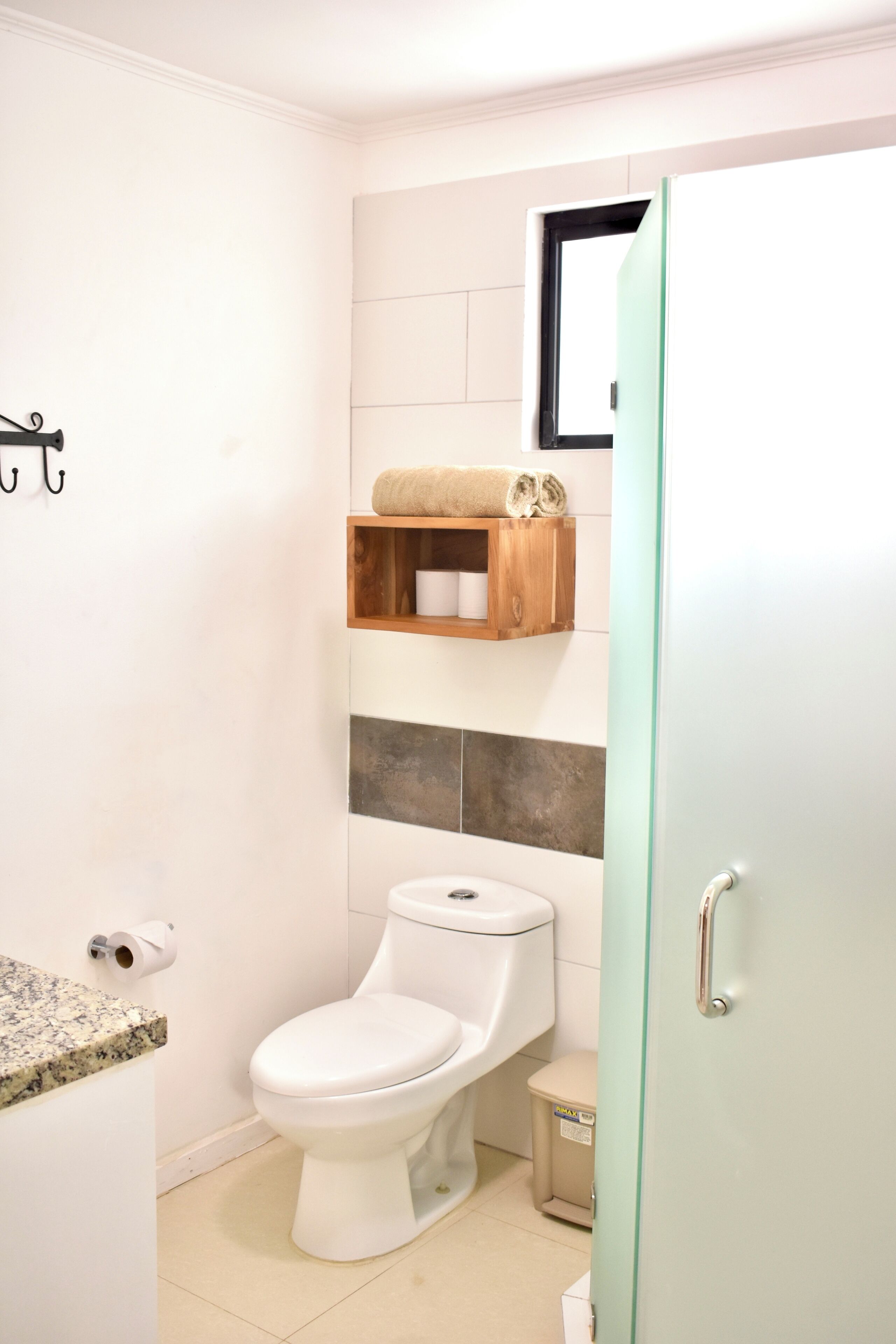 Un baño con inodoro, un pequeño bote de basura, una estantería de madera con toallas y un dispensador de papel higiénico.
