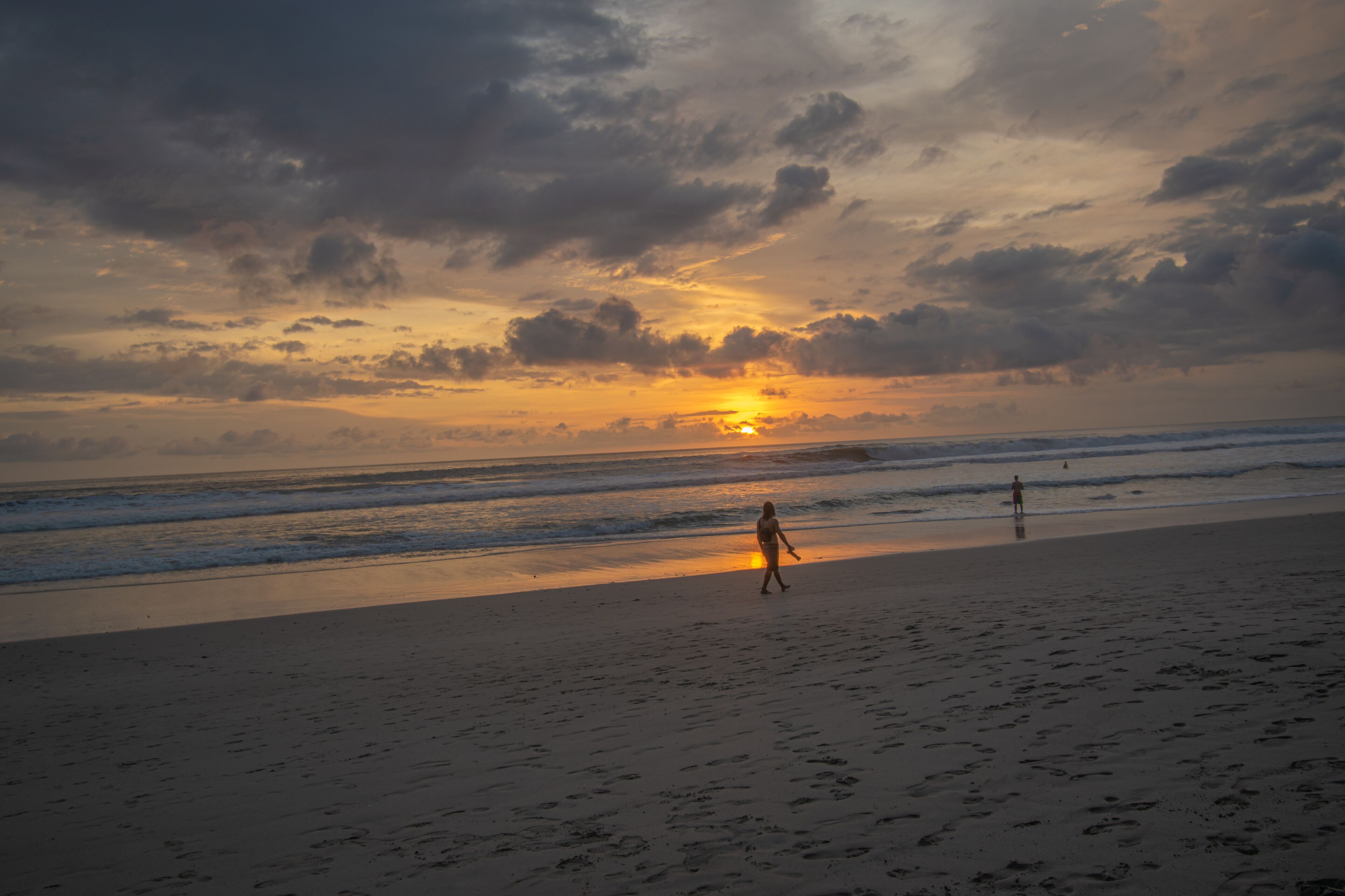 Escena de playa con huellas que conducen al agua, una persona caminando y el sol poniéndose en el horizonte.