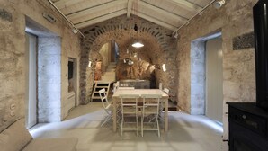 Une pièce rustique aux murs de pierre, avec une table et des chaises en bois, une cheminée en pierre et un poêle noir.
